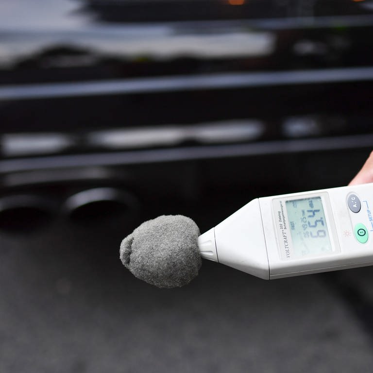 Dezibel-Messgerät vor einem Autoauspuff