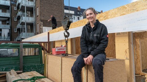 Eine junge, blonde Frau sitzt lächelnd in Arbeitskleidung auf einem Stapel OSB-Platten auf einer Baustelle. (Foto: SWR)