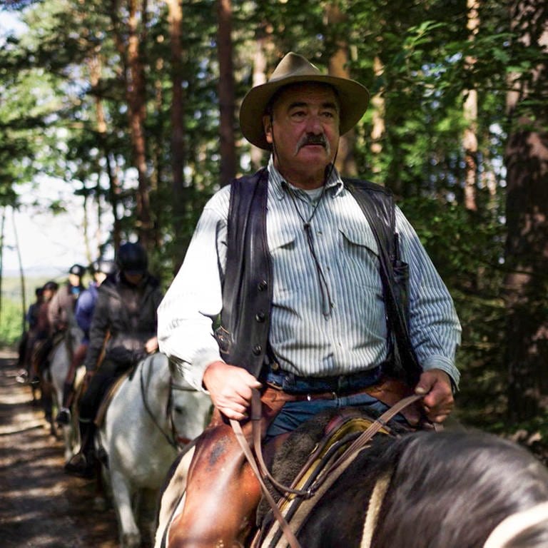 Mann mit Cowboyhut und Schnurrbart sitzt auf Pferd, hinter ihm andere Reiter.