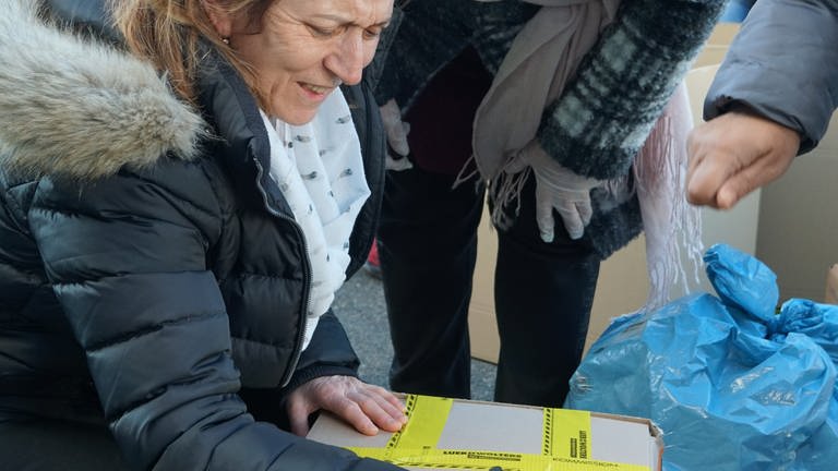 Ältere Frau, in schwarzer Jacke mit weißem Schal kniet vor einem Karton und beschriftet ihn.  (Foto: SWR)