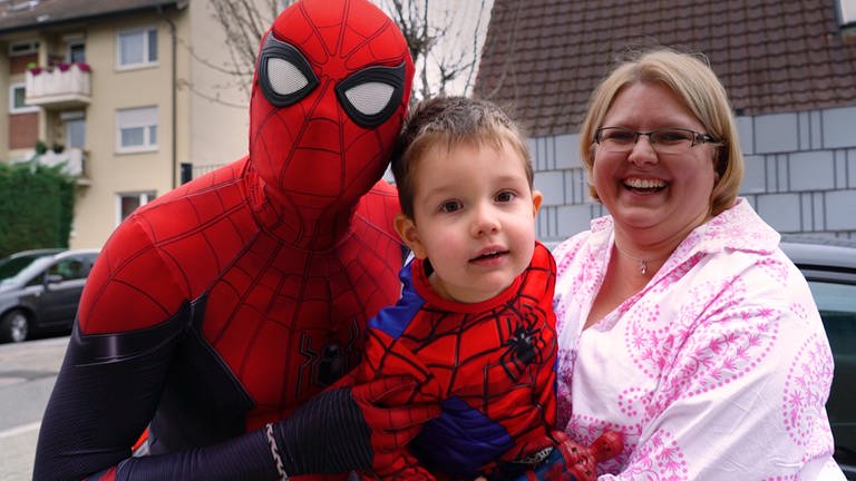 Drei Personen lächeln in die Kamera: Ein Mann als Spiderman verkleidet, ein Kleinkind und seine Mutter