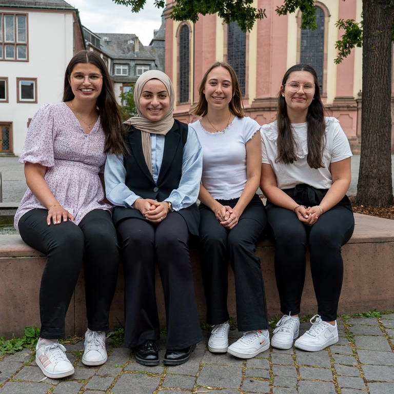 Vier junge Finanzbeamtinnen sitzen nebeneinander auf dem Rand eines Brunnens auf dem Wormser Marktplatz und lächeln.