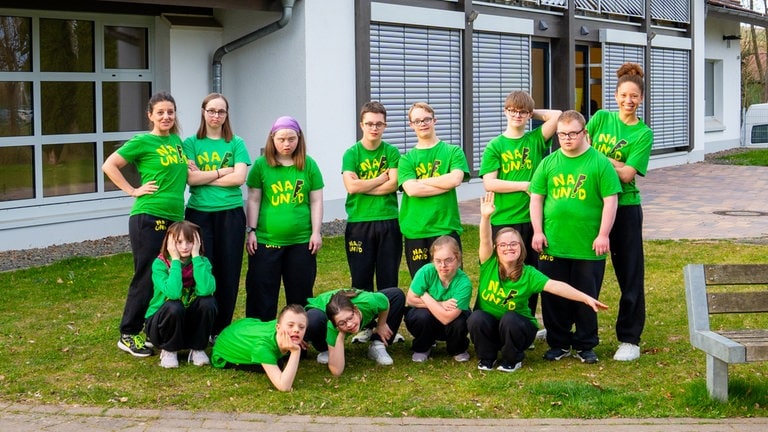 Eine Gruppe Jugendlicher mit Downsyndrom steht mit zwei erwachsenen Frauen auf einer Wiese und lächeln. Alle tragen grüne T-Shirts. (Foto: SWR) Eine Gruppe Jugendlicher mit Downsyndrom steht mit zwei erwachsenen Frauen auf einer Wiese und lächeln. Alle tragen grüne T-Shirts.