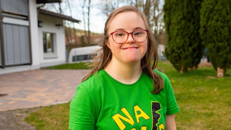 Ein Mädchen mit blonden Haaren und Downsyndrom lächelt in die Kamera. Sie trägt ein grünes T-Shirt und eine Brille. (Foto: SWR) Ein Mädchen mit blonden Haaren und Downsyndrom lächelt in die Kamera. Sie trägt ein grünes T-Shirt und eine Brille.