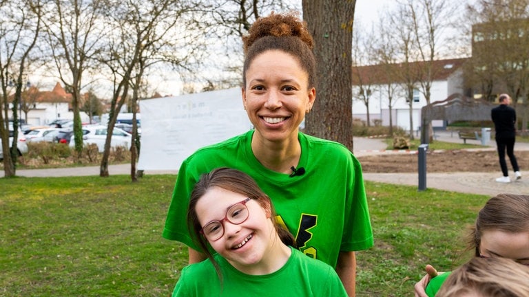 Eine Frau mit braunen Haaren und grünem T-Shirt lächelt in Kamera, vor ihr ein Mädchen mit Downsyndrom und grünem T-Shirt, das lächelt. (Foto: SWR) Eine Frau mit braunen Haaren und grünem T-Shirt lächelt in Kamera, vor ihr ein Mädchen mit Downsyndrom und grünem T-Shirt, das lächelt.
