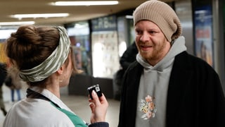 Dominik steht in einer Unterführung, trägt eine beigefarbene Mütze, einen hellen Pullover und darüber einen schwarzen Mantel. Er lächelt.