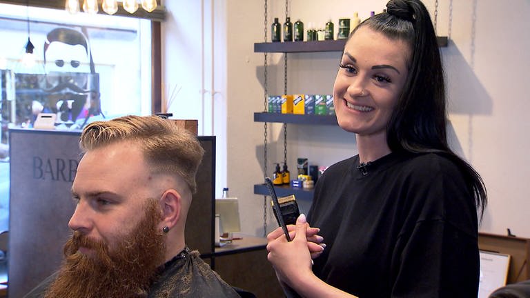 Friseurmeisterin und Barberin Vanessa schneidet einem Kunden in ihrem eigenen Laden die Haare und lächelt dabei in die Kamera.