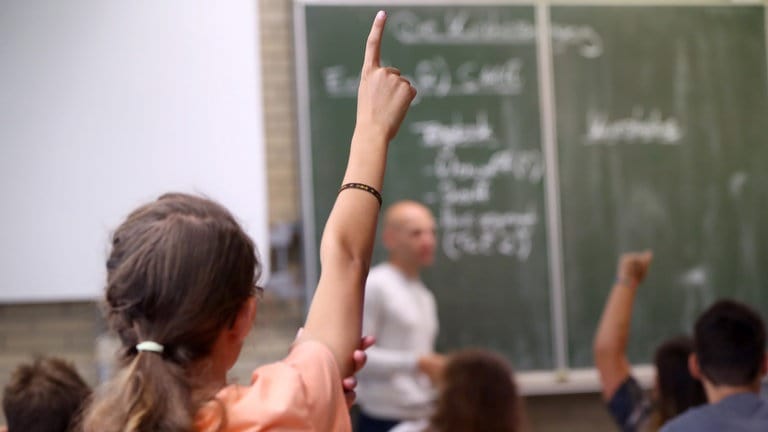 Ein Mädchen streckt im Unterricht den Finger, ein Lehrer erklärt an der Tafel: Demokratiebildung ist Aufgabe von Schule. Der sogenannte "Beutelsbacher Konsens" aus dem Jahr 1976 legt die Prinzipien dafür fest: Keine Indoktrination im Klassenzimmer, politische Kontroversen sind auch kontrovers darzustellen, zudem sollen Schüler*innen befähigt werden, sich eine eigene Meinung zu bilden.  (Foto: dpa Bildfunk, picture alliance/dpa | Daniel Karmann)