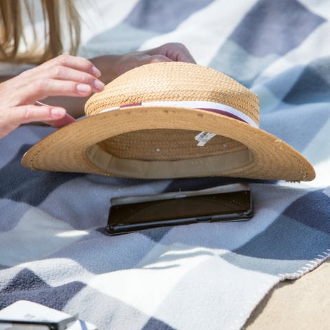 Ein auf einer Decke liegendes Handy wird mit einem Sonnenhut vor Sonne geschützt.  (Foto: picture-alliance / Reportdienste, picture alliance / dpa-tmn | Christin Klose)