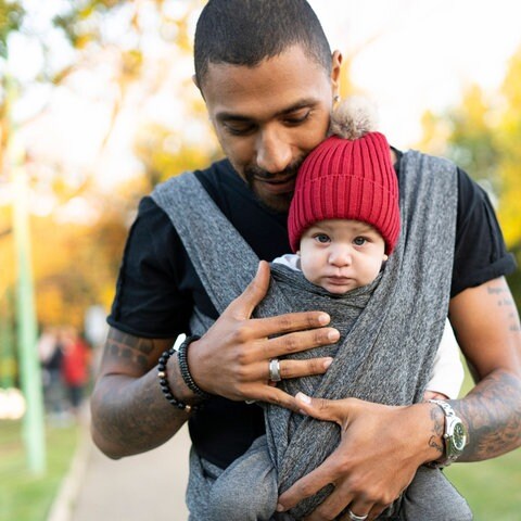 Vater hält schützend seine Hande um das Baby, das er im Tragetuch vor sich hält: Heute möchten immer mehr junge Männer liebevolle und präsente Väter sein. Und auch die Forschung ergründet immer besser, was Männer im Umgang mit Kindern oft anders machen. Klar ist: Kinder können in ihrer Entwicklung sehr von aktiven Vätern profitieren. (Foto: IMAGO, IMAGO / Westend61)