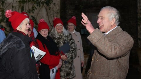 Rechts steht ein älterer Mann im Mantel mit erhobener Hand und offenem Mund, als würde er was sagen und dirigieren, ihm gegenüber stehen meherere Frauen mit roten Wollpudelmützen - König Charles und die Sternsinger (Foto: dpa Bildfunk, Picture Alliance)