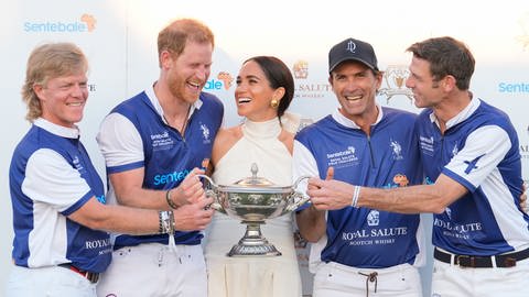 Prinz Harry im Trikot samt seinem Polo-Team und in der Mitte mit Meghan in einem weißen Kleid halten gemeinsam die Trophäe nach einem gewonnen Spiel in Florida. (Foto: picture-alliance / Reportdienste, picture alliance/dpa/AP | Rebecca Blackwell) Prinz Harry im Trikot samt seinem Polo-Team und in der Mitte mit Meghan in einem weißen Kleid halten gemeinsam die Trophäe nach einem gewonnen Spiel in Florida.