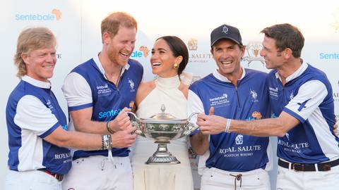 Prinz Harry im Trikot samt seinem Polo-Team und in der Mitte mit Meghan in einem weißen Kleid halten gemeinsam die Trophäe nach einem gewonnen Spiel in Florida. (Foto: picture-alliance / Reportdienste, picture alliance/dpa/AP | Rebecca Blackwell)