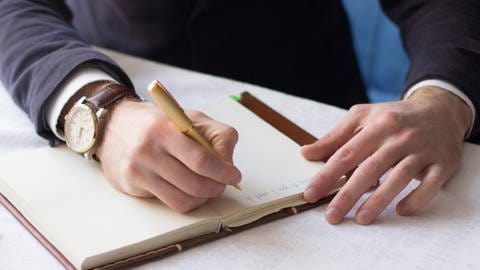 Dankbarkeitstagebuch: Man sieht ein leeres offenes Buch mit weißen Seiten und zwei Männerhände. An der einen Hand trägt der Mann eine Armbanduhr und er hält damit einen Stift in der Hand, mit dem er etwas auf das Papier schreibt. 