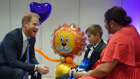 Prinz Harry sitzt einer Mutter und ihrem Sohn gegenüber. Sie plaudern und der Royal lacht dabei. Im Hintergrund sind bunte Luftballons zu sehen. (Foto: dpa Bildfunk, Picture Alliance)