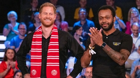 Prinz Harry lacht freudig. Er trägt ein schwarzes Hemd und um den Hals einen Mainz 05 Schal im aktuellen Sportstudio des ZDF. Neben ihm steht Angelo Anderson, der für die "Invictus Games Foundation" arbeitet. (Foto: picture-alliance / Reportdienste, picture alliance/dpa/ZDF | Ralph Orlowski)