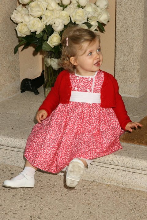 Die spanische Prinzessin Leonor sitzt als kleines Mädchen von einem Jahr in einem rotgeblümten Kleidchen und mit blonden Locken auf einer Treppenstufe vor einer Vase mit weißen Rosen und schaut zur Seite weg. (Foto: IMAGO, IMAGO / CordonPress)