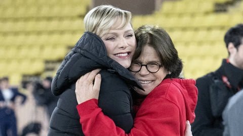 Fürstin Charlène von Monaco und ihre Schwägerin Stéphanie von Monaco umarmen sich ganz innig uns strahlen dabei in die Kamera. Charlène trägt einen schwarzen dicken Wintermantel, Stéphanie einen roten Kapuzenpullover. Sie stehen im Stadion von Monaco auf dem Spielfeld, im Hintergrund sieht man Männer auf dem Spielfeld und die leeren gelben Sitzplätze. (Foto: picture-alliance / Reportdienste, picture alliance / abaca | Niviere David/ABACAPRESS.COM) Fürstin Charlène von Monaco und ihre Schwägerin Stéphanie von Monaco umarmen sich ganz innig uns strahlen dabei in die Kamera. Charlène trägt einen schwarzen dicken Wintermantel, Stéphanie einen roten Kapuzenpullover. Sie stehen im Stadion von Monaco auf dem Spielfeld, im Hintergrund sieht man Männer auf dem Spielfeld und die leeren gelben Sitzplätze.