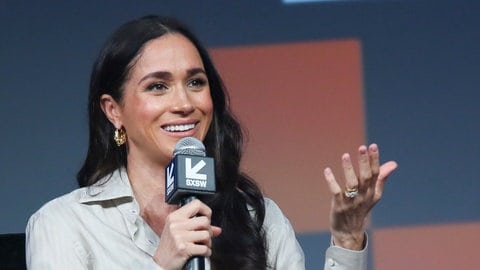 Meghan Markle trägt ihr schwarzes Haar offen und gewellt, ihre goldenen Ohrringe glänzen und sie trägt ein helles Oberteil, das aussieht wie eine Bluse mit Kragen. Die obersten Knöpfe davon sind geöffnet. In der rechten Hand hält sie ein Mikrofon in das sie spricht. Sie lächelt. (Foto: picture-alliance / Reportdienste, picture alliance/dpa/Invision via AP | Jack Plunkett) Meghan Markle trägt ihr schwarzes Haar offen und gewellt, ihre goldenen Ohrringe glänzen und sie trägt ein helles Oberteil, das aussieht wie eine Bluse mit Kragen. Die obersten Knöpfe davon sind geöffnet. In der rechten Hand hält sie ein Mikrofon in das sie spricht. Sie lächelt.
