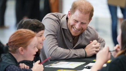 Prinz Harry sitzt an einem Tisch mit Kindern. Er trägt ein hellbraunes Hemd und beugt sich zu den Kindern nach unten und lächelt. (Foto: picture-alliance / Reportdienste, picture alliance/dpa/Canadian Press via ZUMA Press | Darryl Dyck) Prinz Harry sitzt an einem Tisch mit Kindern. Er trägt ein hellbraunes Hemd und beugt sich zu den Kindern nach unten und lächelt.