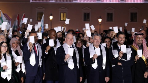 Ein Gruppenbild mit Albert, dem Fürsten von Monaco, der im schwarzen Anzug in der Mitte steht, alle Leute auf dem Bild halten einen weißen Zettel hoch und tragen eine weiße Stola um den Hals (Foto: dpa Bildfunk, Picture Alliance) Ein Gruppenbild mit Albert, dem Fürsten von Monaco, der im schwarzen Anzug in der Mitte steht, alle Leute auf dem Bild halten einen weißen Zettel hoch und tragen eine weiße Stola um den Hals