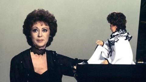 Caterina Valente sitzt am Klavier, darauf eine Puppe. Bild aus der Sendung "Wünsche, die ich mir erfüllte", Januar 1981. (Foto: SWR, SWR/Eraki Entertainment - Eraki Entertainment)