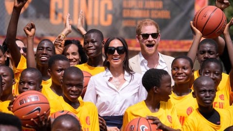 Prinz Harry und Herzogin Meghan stehen in einer Traube Jugendlicher und lachen in die Kamera. (Foto: dpa Bildfunk, picture alliance/dpa/AP | Sunday Alamba) Prinz Harry und Herzogin Meghan stehen in einer Traube Jugendlicher und lachen in die Kamera.