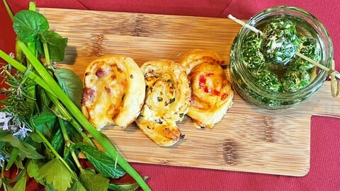 drei Blätterteigschnecken mit verschiedenem Frischkäse auf einem Holzbrett, daneben ein Glas mit Rucola-Frischkäsekugeln und ein Bündel Kräuter (Foto: SWR, Alexandra Daub)