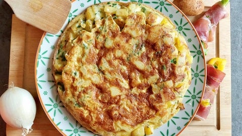 Tortilla auf einem hellen Teller, daneben eine Zwiebel, ein Holzschaber und Gemüse in Schinken gewickelt (Foto: SWR, Jens Alinia und Corinne Schied)