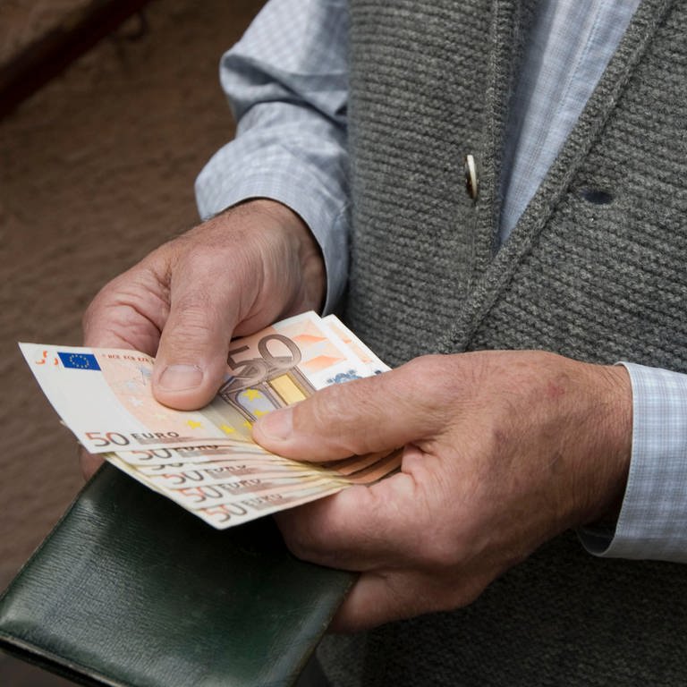 Ein älterer Mann mit Strickweste hält mehrere 50-Euro-Scheine aufgefächert in der Hand