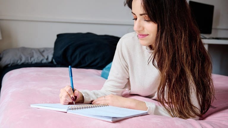 Dankbarkeitstagebuch: Eine Frau mit langen dunklen Haaren liegt im Bett und schreibt in ein leeres Buch mit einem blauen Stift. Die positiven Gedanken dabei scheinen sie zum Lächeln zu bewegen und ihrer Stimmung gut zu tun.