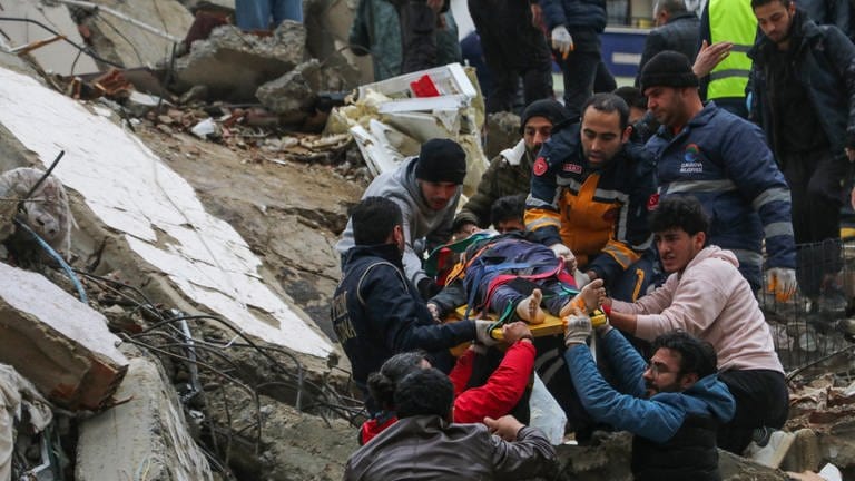 Menschen und Rettungskräfte bergen eine Person auf einer Bahre aus einem eingestürzten Gebäude.