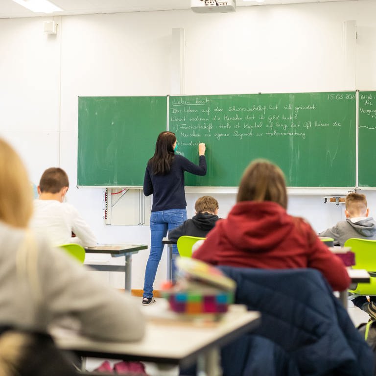 Eine Lehrerin steht an einer Tafel in einem Klassenzimmer, Schüler lernen. Ab dem kommenden Wintersemester können Studierende in einem Modellversuch in einem dualen Masterstudiengang auf Lehramt studieren. 
