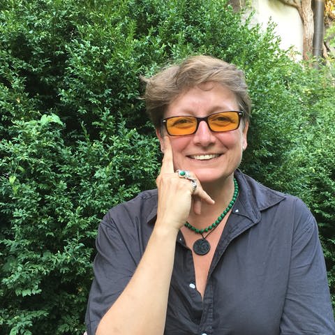 Frau mit gelb getönter Brille blickt in die Kamera, im Hintergrund grünes Gebüsch (Foto: SWR, Esther Leuffen)