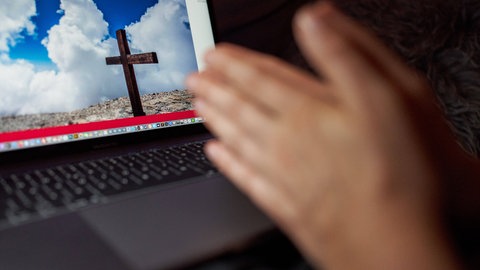 Symbolbild: Auf einem Computerbildschirm ist ein Kreuz zu sehen, davor Hände (Foto: IMAGO, IMAGO / Action Pictures)