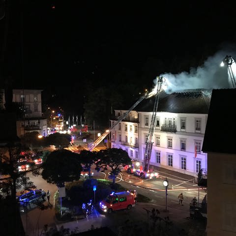 Aus dem Dachstuhl eines Hotels in Baden-Baden dringen weiße Rauchschwaden, während die Feuerwehr den Brand von Drehleitern aus bekämpft.