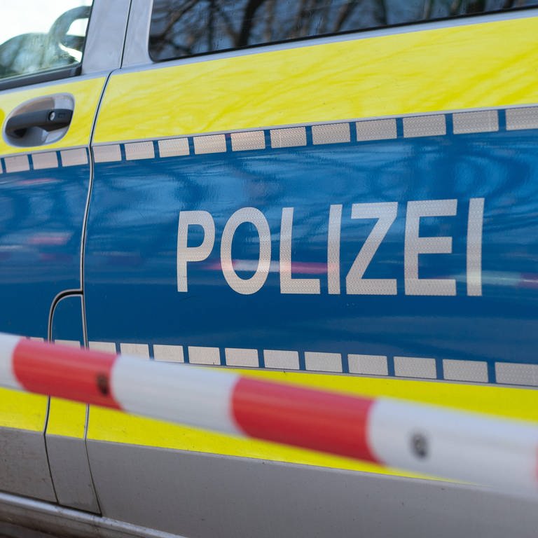 Absperrband vor einem Polizeiauto. Tötungsdelikt in Waldbronn: Nachdem eine 69-jährige Frau tot in ihrer Wohnung gefunden wurde, ist nun bekannt, dass ihr Sohn als tatverdächig gilt.