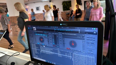 Ein Monitor zeigt abgespielte Musik, im Hintergrund tanzen Menschen