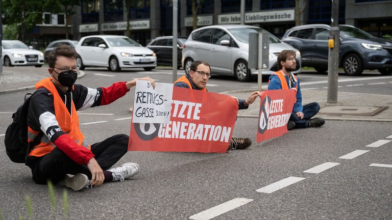 Drei Klimaaktivisten der Gruppe "Letzte Generation" blockieren eine Straße in Stuttgart. 