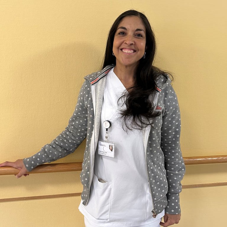 Anita Singh-Carl steht in einem weißen Krankenpflegerinnen-Outfit vor einer gelben Wand in der Uniklinik.
