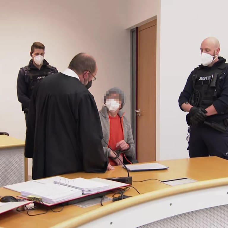 Der mutmaßlicher "Reichsbürger" sitzt zu Prozessbeginn im Gerichtssaal des Oberlandesgerichts in Stuttgart auf einem Stuhl. Er trägt einen orangenen Pullover, darüber eine graue Jacke. Neben ihm steht sein Verteidiger, der trägt die schwarze Anwaltsrobe. Neben den beiden stehen zwei Polizisten. Alle tragen FFP2-Masken. (Foto: SWR)