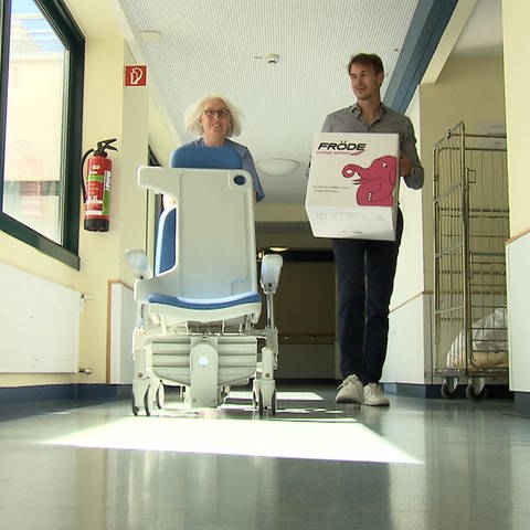 Zwei Mitarbeitende im Krankenhausflur beim Umzug, mit Kiste und medizinischem Stuhl (Foto: SWR)
