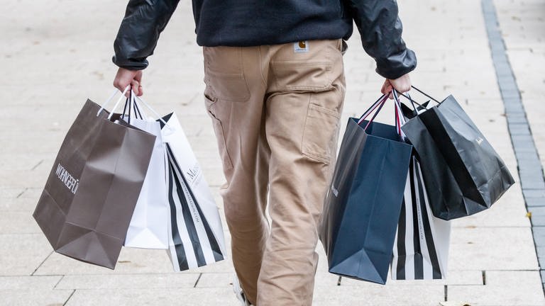 Ein Mann trägt mehrere Einkaufstüten von einem erfolgreichen Shoppingtag, Einkaufstag. Von der Person ist die untere Körperhälfte von hinten zu sehen.