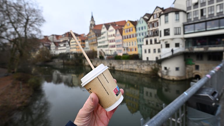 Eine Hand hält einen Einweg-Kaffeebecher vor dem Hintergrund der Tübinger Altstadt (gestellte Szene).
