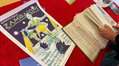 Dreifarbig gedrucktes Plakat aus dem Eröffnungsjahr des Zirkus Zamboioni. Auf dem Siebdruckbild in lila, gelb und rosa ist eine Seiltänzerin im Tutu abgebildet. 