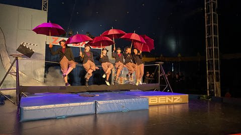 Sechs junge Frauen mit roten Regenschirmen sitzen aufgereiht auf einem Balancier-Seil