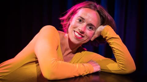 Bekleidet mit einem gelben Shirt lächelt die Kleinkünstlerin Judith Bach beleuchtet vom Bühnenlicht in die Kamera. 