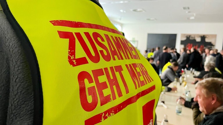 Eine gelbe Streikweste im Vordergrund. Menschen sitzen an einem Tisch.  (Foto: SWR, Anja Müller)