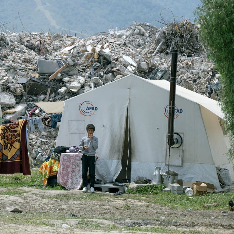 Junge steht vor einem Notunterkunft-Zelt, hinter ihm Berge von Schutt: Die Stadt Aalen (Ostalbkreis) will den Bau einer Rehaklinik in der türkischen Partnerstadt Antakya mitfinanzieren, die vor einem Jahr von einem Erdbeben betroffen war.