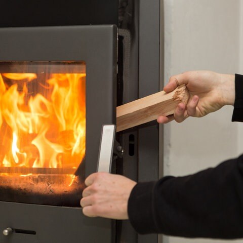 Ein Mann legt in seiner Wohnung ein Stück Holz in einen Kaminofen, in dem ein Feuer brennt. (Foto: dpa Bildfunk, Picture Alliance)